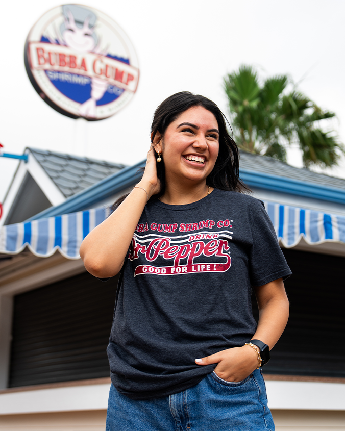 A young woman in a dark t-shirt with "Bubba Gump Shrimp Co." and "Dr Pepper" printed on it, standing in front of a restaurant.