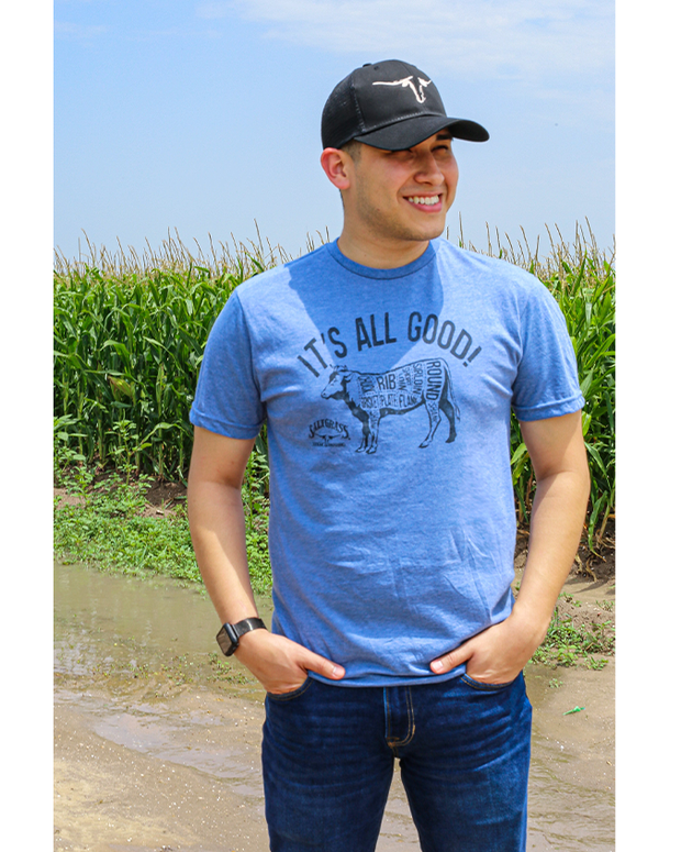 man in crop field wearing black hat and tee. Indigo tee with "It's All Good!" and cow silhouette with word cloud of meat that comes from cow.