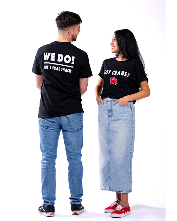 Two individuals standing side by side against a white background. They are both wearing black t-shirts with white text and logos on the back. The person on the left is dressed in blue jeans and sneakers, and their shirt has the text “WE DO! JOE’S CRAB SHACK.” The person on the right is wearing light blue denim jeans and red sneakers, and their shirt reads “GOT CRABS?” with a crab logo below. Their relaxed stance allows the back of their shirts, highlighting the printed text and logos, to be clearly visible.