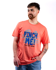 The image shows a person standing against a white background, wearing coral t-shirt with the text “PINCH ME!” in large blue letters and “JOE'S CRAB SHACK” in smaller blue letters below. Surrounding the text are graphics of crab claws.