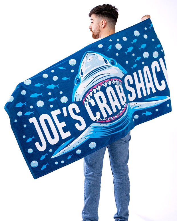 male model, holding towel open on him back. Blue towel that has bubble and fish swimming away from shark. in center is a great white shark with its mouth open, crunching onto word "crab". across the towel, going through the shark's mouth, reads in white letters "joe's crab shack". 