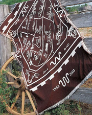 Rectangular throw blanket with fringed edges. The blanket features a maroon and white color scheme and a central design that includes various cattle, horses and cowboys-themed graphics. King Ranch icon logo with the text 'EST. 1853' at the bottom. Throw blanket laying on a side of a wood fence and a  wood carriage wheel giving it a feel like at a farm.
