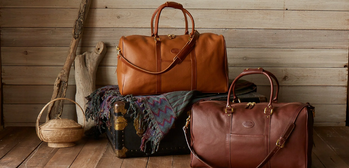 The image features two large leather bags against a rustic wooden plank background, evoking a sense of travel and adventure with a vintage flair. A woven basket and a multicolored textile add to the scene’s charm.