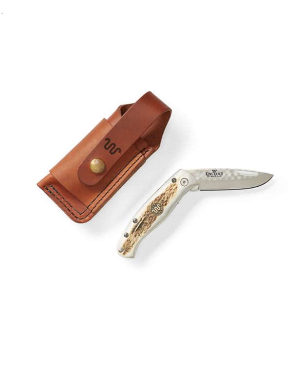 folding pocket knife with a decorative handle featuring King Ranch logo on the blade and handle, next to the knife is a brown leather sheath. The sheath has a snap button closure and is embossed with King Ranch logo. Items displayed on a white background.