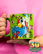 A vibrant green mug adorned with a colorful illustration of a parrot surrounded by tropical flowers, held against a pink shirt. The mug features an emblem celebrating ‘30 WILD YEARS!’ of Rainforest Cafe with Cha Cha Sticking out the 0.