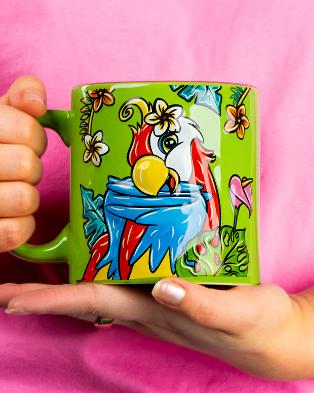 A close-up view of a model in a pink shirts' hands holding a colorful green mug with a posing parrot surrounded by tropical flowers and leaves.