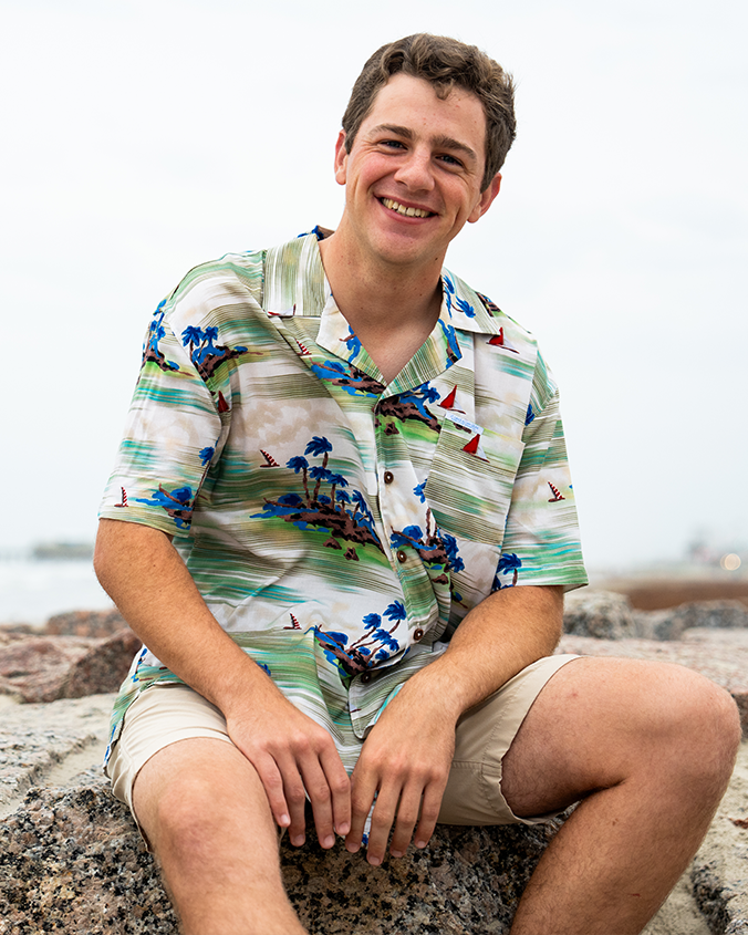A person in a colorful tropical shirt sits on a rocky shore, with a scenic beach and cloudy sky in the background.
