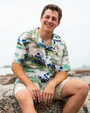 A person in a colorful tropical shirt sits on a rocky shore, with a scenic beach and cloudy sky in the background.