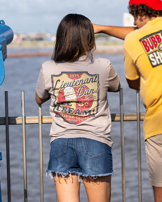 girl with guy leaning on railing facing the beach. she is wearing denim shorts and Beige short sleeve t-shirt with retro graphics on the back, resembling a rectangular vintage sign in muted mustard and red colors. The sign features an ice cream cone with the price of "5c" and the text "Lieutenant Dan" on top.