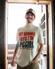 A waist-up view of a tattooed model standing in front of an open wooden door with his hand in his jean pocket, wearing a cream-colored shirt that reads "MAMA SAYS I'M SPECIAL, BUBBA GUMP SHRIMP CO." in the center. He is also brown and white baseball cap with the same wording. 