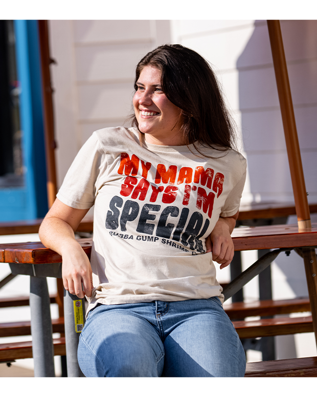 female model leaning against picnic table wearing Sand ringspun cotton tee with "My mama says I'm special" in orange ombre retro font and "Bubba Gump Shrimp Co" underneath it.