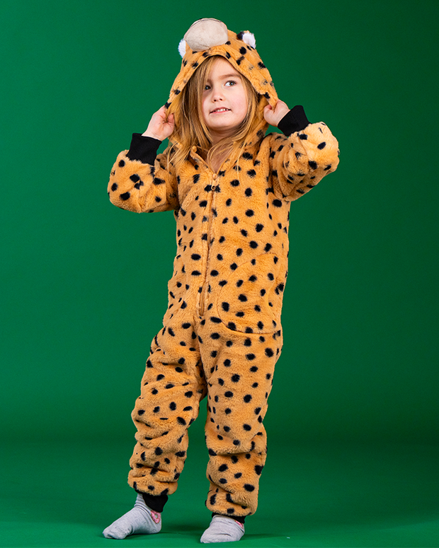 A kid wearing a cheetah-print onesie with ears and a tail, standing against a green background. They are adjusting the hood with ears attached to it.