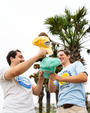 Three friends hold colorful hats, surrounded by palm trees and a cloudy sky, enjoying a casual moment outdoors.