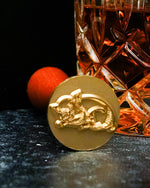 A close-up of a gold ice stamp laying on a marble black surface against a black background. It has an intricate pig design and a wooden handle seen at an angle, placed in front of a crystal glass full of liquid. 