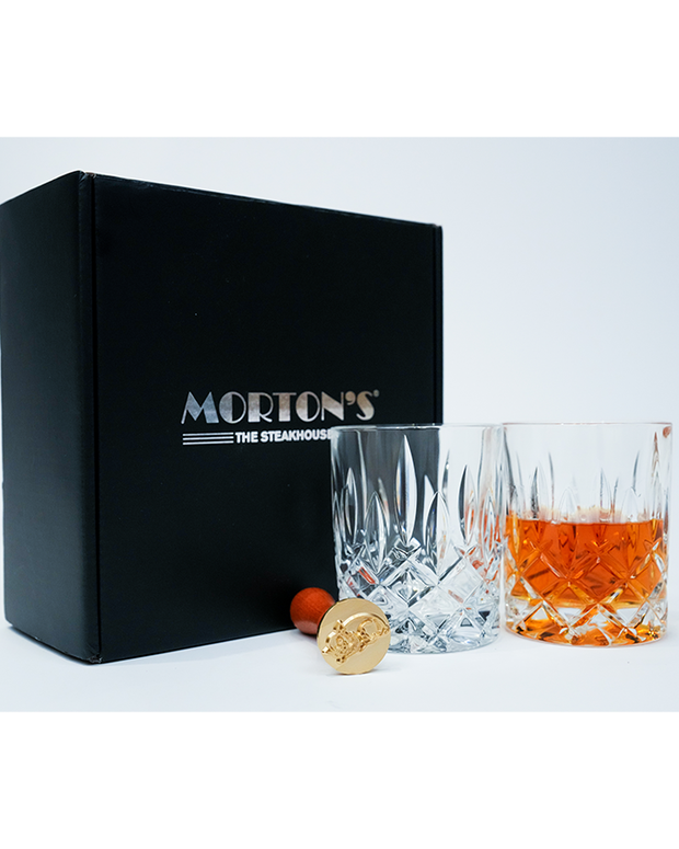A black Morton’s The Steakhouse box with silver lettering is positioned on the left side of the image against a white background. In front of the box, to the right, there are two glasses; one is empty with a cut crystal pattern, and the other contains an amber-colored liquid, presumably a beverage. Next to the glasses you can see the the pig ice stamp.
