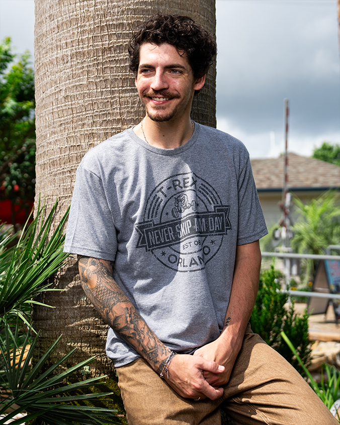 A person in a gray T-shirt with "Never Skip Arm Day" text poses next to a palm tree, showcasing a detailed arm tattoo in a sunny setting.