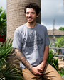 A person in a gray T-shirt with "Never Skip Arm Day" text poses next to a palm tree, showcasing a detailed arm tattoo in a sunny setting.