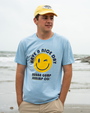 A young man stands on a beach, wearing a light blue shirt with a smiley face and the text "Have a Nice Day, Bubba Gump Shrimp Co."