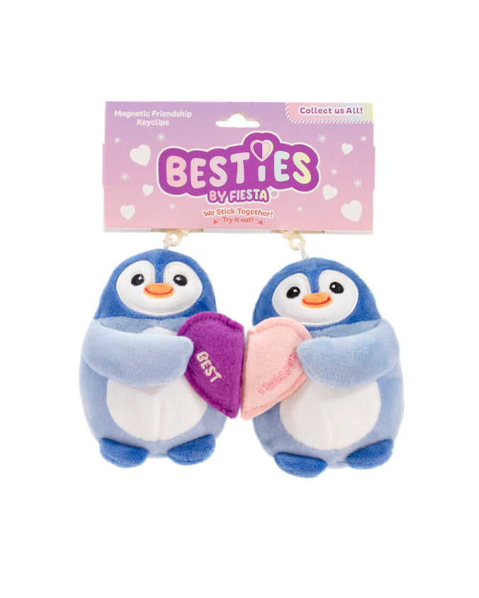 Two plush penguin keychains with magnetic attachments on half hearts they are holding and connects to each other. One says "best" and the other "friends". The penguins are blue and white, and they are suspended in front of a packaging card that says ‘Magnetic Friendship Keyclip’ at the top and ‘BESTIES by Fiesta’ below. The card also has the text ‘Collect us ALL!’ with decorative hearts around it.