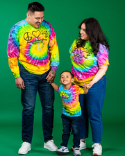 A family of three, wearing colorful tie-dye shirts adorned with peace symbols and hearts, stands together against a green background. The adults hold hands with the child in the center, all sporting dark blue jeans. The text ‘Peace Love Frogs’ ppears on all shirts.