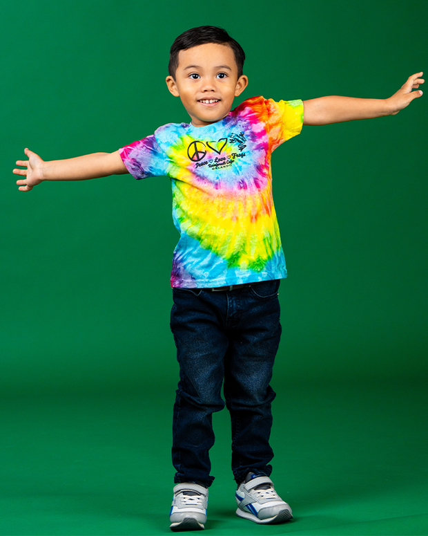 A person stands with arms outstretched against a solid green background, wearing a vibrant tie-dye t-shirt featuring a peace symbol and the text ‘Peace Love Frogs.’ The colorful shirt is matched with dark blue jeans and white sneakers with light blue accents. 