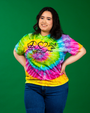 A person stands against a solid green background, wearing a vibrant tie-dye t-shirt bursting with a swirl of bright colors including yellow, green, blue, pink, and purple. The shirt is adorned with symbols such as a peace sign, frog and a heart, and the text ‘Peace Love Frogs Rainforest Cafe .’ The individual’s hands are on their hips, and they are dressed in dark blue jeans.
