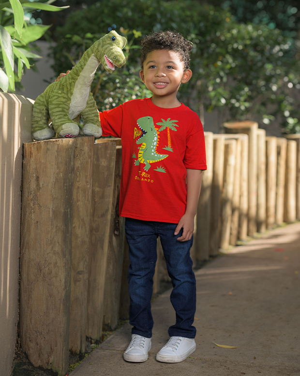 A kid stands outdoors next to a wooden fence, wearing a red t-shirt featuring a green dinosaur and palm tree design, paired with dark blue jeans and white sneakers. A plush green dinosaur toy is perched on the fence beside them. The setting is lush with greenery and bathed in natural sunlight.