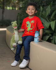 A kid sits on a beige concrete ledge, holding a green dinosaur plush toy in their left hand and a blue water bottle in their right. They are dressed in a red t-shirt with a dinosaur graphic and the text ‘T-Rex Orlando,’ paired with dark blue jeans and white sneakers. They are surrounded by lush green plants.