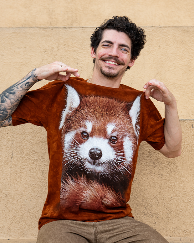 A person displays a brown t-shirt featuring a detailed print of a red panda's face against a textured beige background.