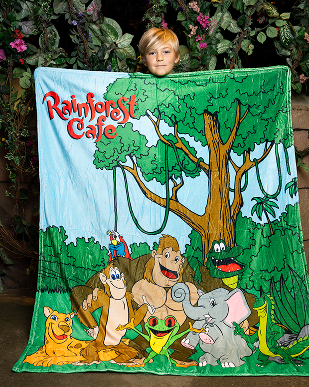 A colorful Rainforest Cafe blanket featuring cheerful animal characters like a monkey, elephant, and parrot against a lush green backdrop.