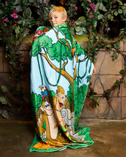 A child wrapped in a colorful blanket featuring cartoon animals in a jungle scene, surrounded by green plants and vines.