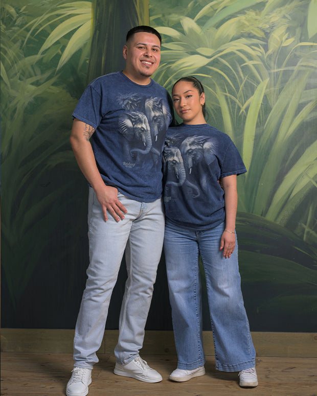 Two individuals pose in front of a backdrop with large green leaves, wearing matching blue t-shirts adorned with an elephant graphic, paired with light-colored jeans and white shoes.