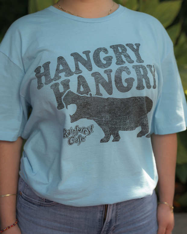 close up of light blue t-shirt with the text ‘HANGRY HANGRY’ and an image of a hippopotamus. The person stands in front of green foliage.