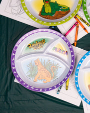 Three similarly designed plates placed in a triangle formation against a table with coloring pages and crayons, with the focus on one plate. The plates are children’s divided plates, the center with three asymmetrical compartments, featuring graphics of a cartoon jaguar against foliage, the "RAINFOREST CAFE" logo, and "MAYA", in the smaller segments, and a purple outline with jungle animal silhouettes.