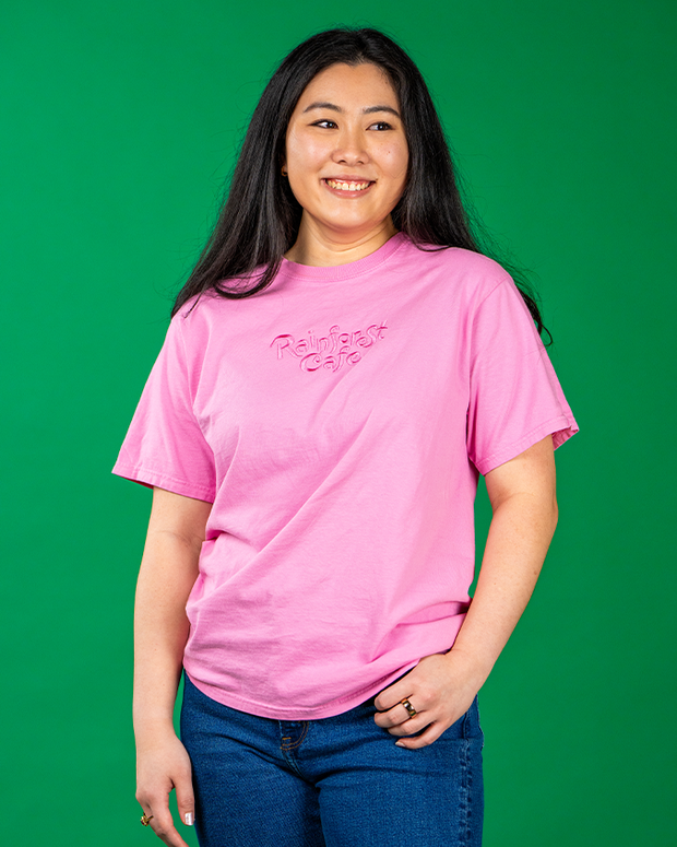 “A person stands against a vibrant green background, wearing a bright pink t-shirt with ‘Rainforest Cafe’ written in white cursive lettering. The individual  has one hand resting on their hip, paired with blue jeans.