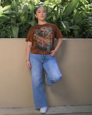 A person is standing against a beige wall, wearing a brown t-shirt adorned with a vibrant print of wildlife animals, paired with light blue jeans and white sneakers. Their right arm is casually resting on the wall, and lush green plants peek over the top of the wall in the background. 