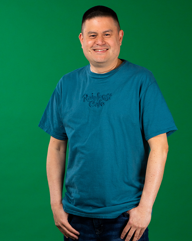 A person in a teal t-shirt with ‘Rainforest Cafe’ in cursive embroidered text stands against a green backdrop, and hands partially in the pockets of their dark pants, exuding a relaxed demeanor.