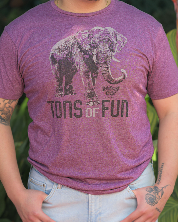 The image depicts a person wearing a purple t-shirt with a playful elephant graphic and the phrase “TONS OF FUN” printed on it. Below the graphic, there’s additional text that reads “Rainforest Cafe.” The individual is also wearing light blue denim jeans and has a tattoo on their left forearm.