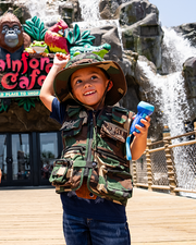 little boy holding a flashlight, standing in front of rainforest cafe sign. he is wearing camo ranger hat and vest with navy top and denim pants.