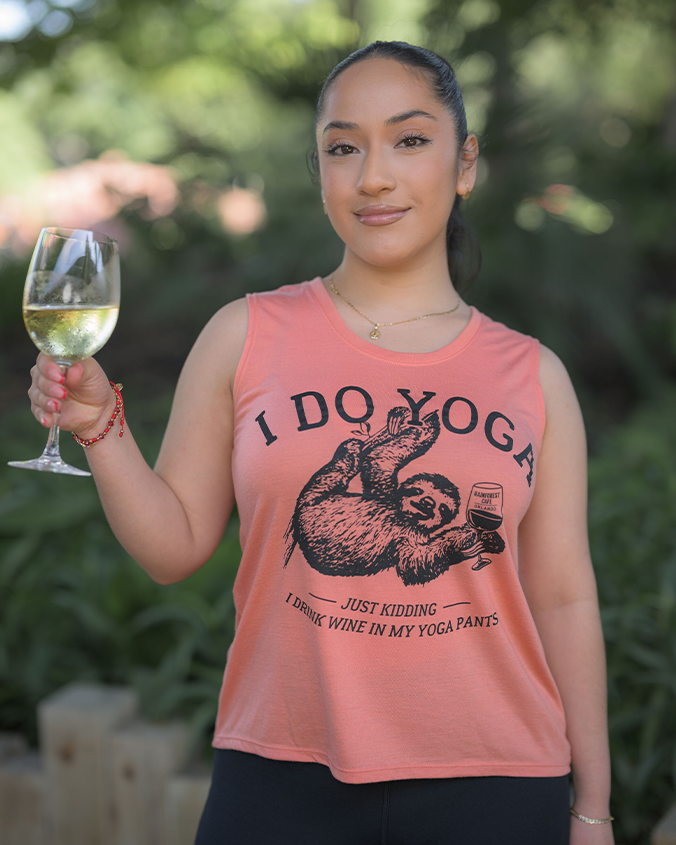 The image features a person standing outdoors holding a glass of white wine. They are wearing a peach-colored tank top with a humorous phrase that reads, “I DO YOGA JUST KIDDING I DRINK WINE IN MY YOGA PANTS,” accompanied by an illustration of a sloth in a yoga pose holding a wine glass. The individual’s face is obscured for privacy. The lush green background suggests a garden or park setting, adding a relaxed and cheerful vibe to the scene.