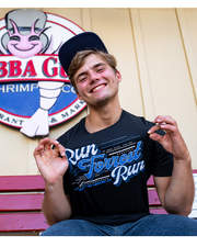 male model wearing Charcoal black cotton tee that says "Run Forrest Run" as a front center chest detail. he is sitting in front of bubba gump logo on a wall and waring a blue cap.