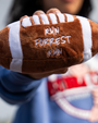 A person holds a plush football featuring the words "RUN FORREST RUN" in white script on a brown, textured surface.
