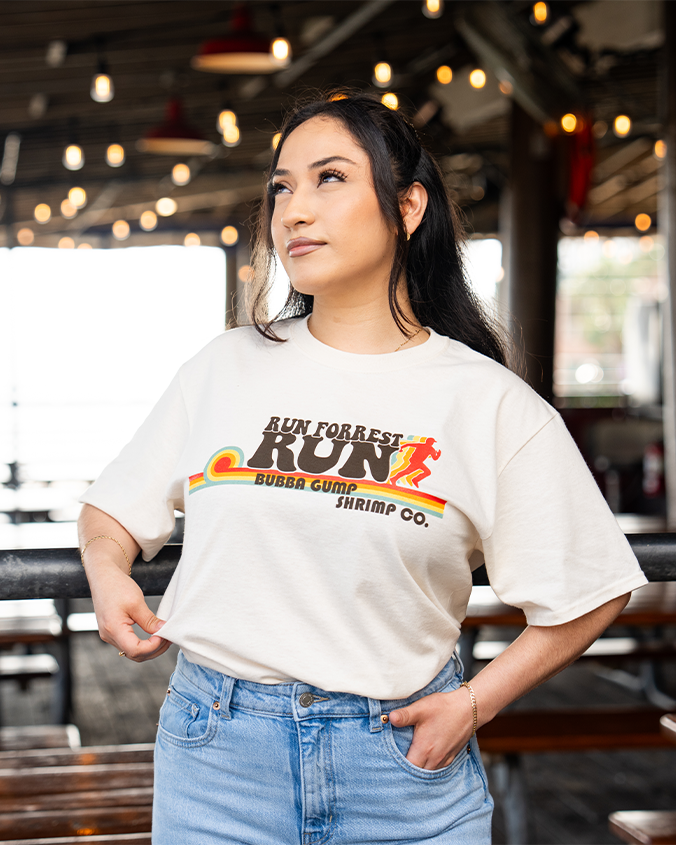 A person in a casual oversized cream t-shirt featuring "RUN FORREST RUN" and a colorful graphic stands in a lively restaurant setting.