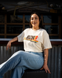A woman sits confidently with a relaxed pose, wearing a beige "Bubba Gump Shrimp Co." t-shirt and light blue jeans, against a rustic background.