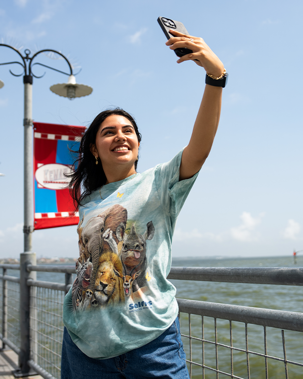A person takes a selfie on a sunny day while wearing a colorful animal-themed T-shirt near a waterfront with decorative lampposts.