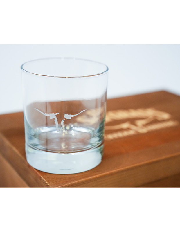 A clear glass featuring an etched design of a longhorn, placed on a wooden surface with partially visible engraved text.