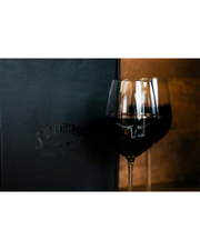 A nearly full glass of deep red wine sits in front of a dark, embossed on a wooden surface, illuminated by soft, ambient lighting, with the word ‘SALTGRASS’ discernible on a black box.