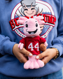 A person holds a pink plush axolotl in a red sports jersey with "44," featuring a smiling face and a small brown football.