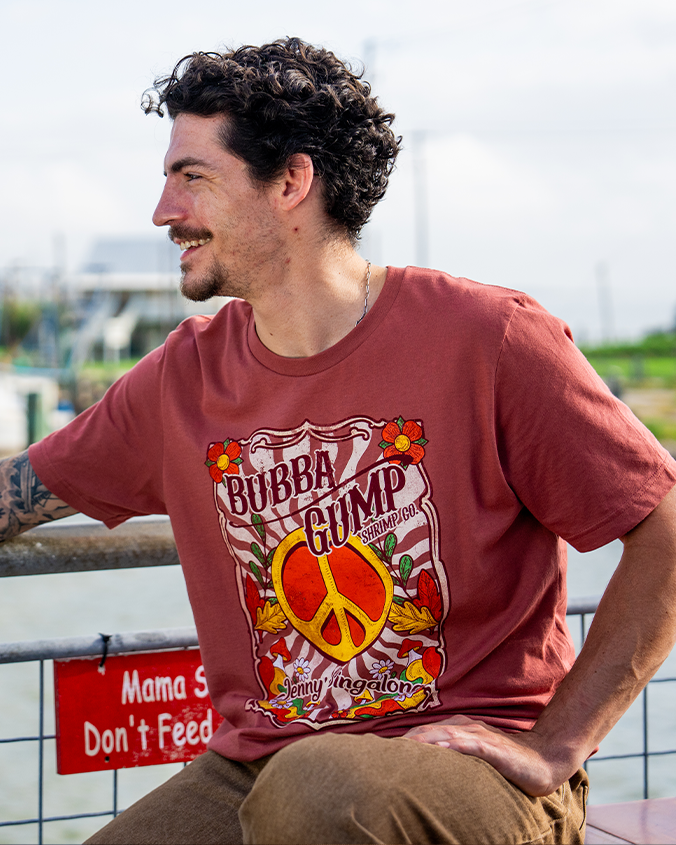A young man sits casually on a railing, wearing a red "Bubba Gump" t-shirt with a peace symbol and floral designs, by a waterway.