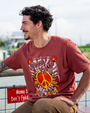 A young man sits casually on a railing, wearing a red "Bubba Gump" t-shirt with a peace symbol and floral designs, by a waterway.